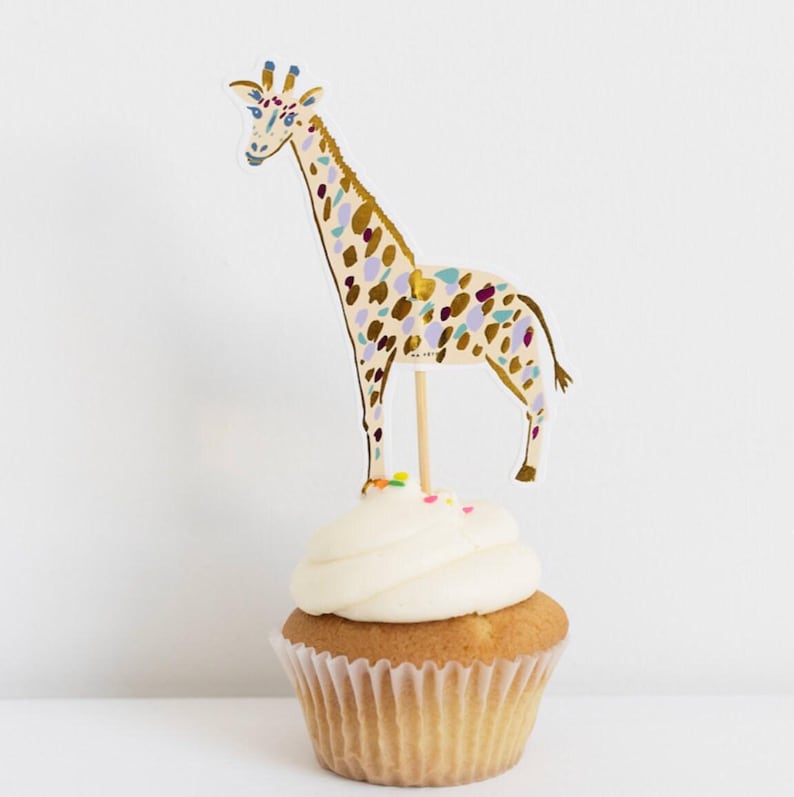 K&ouml;nnte beinhalten: Ein Cupcake mit wei&szlig;em Zuckerguss, verziert mit einem Giraffen-Kuchenaufsatz. Die Giraffe ist beige mit bunten Flecken und einem Holzstab. Der Cupcake befindet sich in einer wei&szlig;en Papierform.