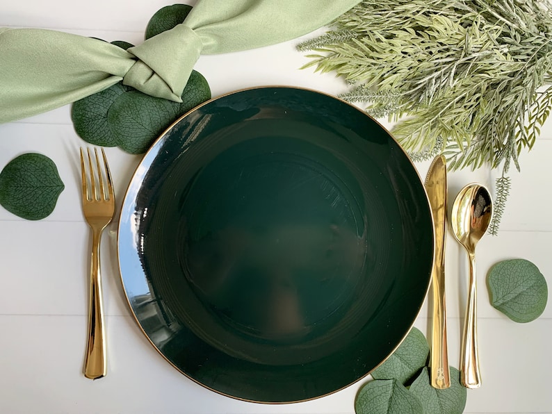Puede incluir: Un plato de cena de color verde oscuro con un borde dorado, un tenedor dorado, un cuchillo dorado y una cuchara dorada. El plato est&aacute; rodeado de hojas verdes.