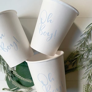 May include: Three white paper cups featuring the text Oh Boy! printed in a light blue script font. The cups are arranged in a cluster, with the text clearly visible on the front of each cup, showcasing a simple design for beverage service.
