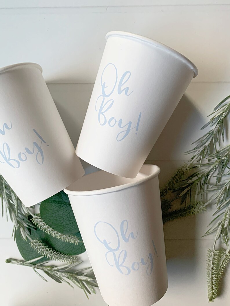 May include: Three white paper cups featuring the text Oh Boy! printed in a light blue script font. The cups are arranged in a cluster, with the text clearly visible on the front of each cup, showcasing a simple design for beverage service.