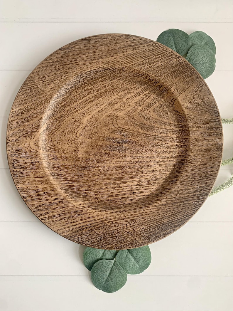May include: A round, wooden charger plate with a dark brown, wood-grain finish. The plate has a slightly raised rim and is surrounded by green leaves. The plate is a decorative item for dining and entertaining.