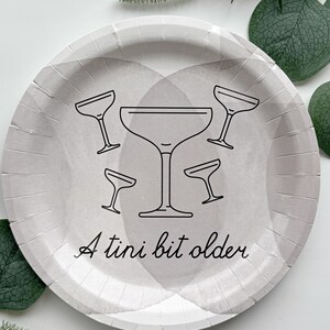 Puede incluir: Un plato redondo de papel gris con un dise&ntilde;o de acuarela y contornos negros de copas de martini. El texto "A tini bit older" est&aacute; escrito en cursiva. La vegetaci&oacute;n decorativa rodea el plato.
