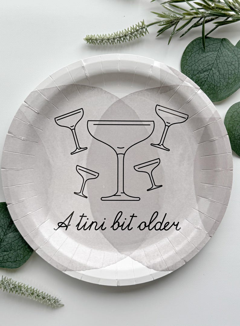Puede incluir: Un plato redondo de papel gris con un dise&ntilde;o de acuarela y contornos negros de copas de martini. El texto "A tini bit older" est&aacute; escrito en cursiva. La vegetaci&oacute;n decorativa rodea el plato.