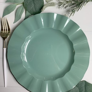 May include: A light green ceramic dinner plate with a ruffled edge. The plate is on a white surface with green leaves.
