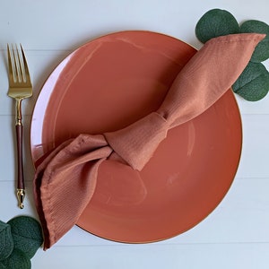 May include: A terracotta-colored dinner plate with a gold rim, a brown napkin folded into a knot, and a gold fork on a white wooden surface.