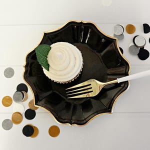 Puede incluir: Un plato de fiesta de papel negro con borde dorado, un solo cupcake con glaseado blanco y una hoja verde en la parte superior, y un tenedor dorado en el plato.