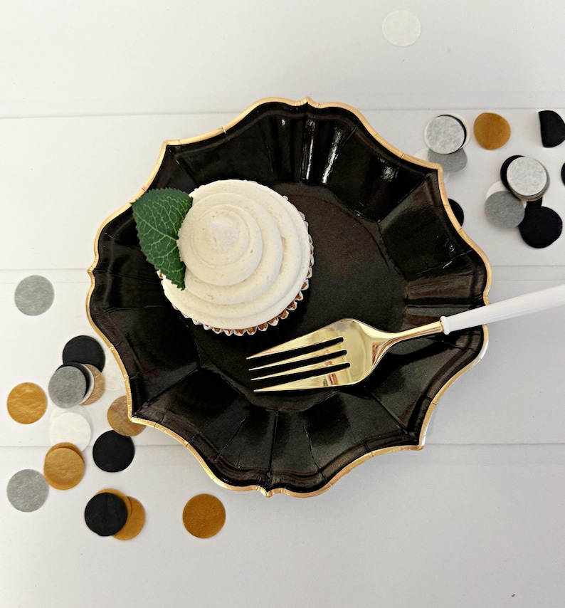 Puede incluir: Un plato de fiesta de papel negro con borde dorado, un solo cupcake con glaseado blanco y una hoja verde en la parte superior, y un tenedor dorado en el plato.