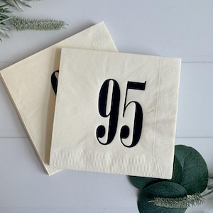 May include: Two cream-colored paper napkins with the number 95 printed in black. The napkins are stacked, with one partially covering the other. Greenery and a white wooden surface are in the background.