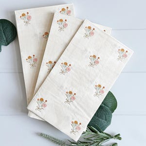 May include: Off-white rectangular napkins with a subtle striped texture. Each napkin is adorned with embroidered floral bouquets in shades of peach, yellow, and pink, tied with a delicate bow. The napkins are arranged in a layered display.