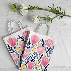 May include: Two white paper gift bags with a floral design featuring pink tulips, navy blue leaves, and yellow accents. The bags have white handles and are arranged on a white wooden surface. A sprig of white flowers is in the background.