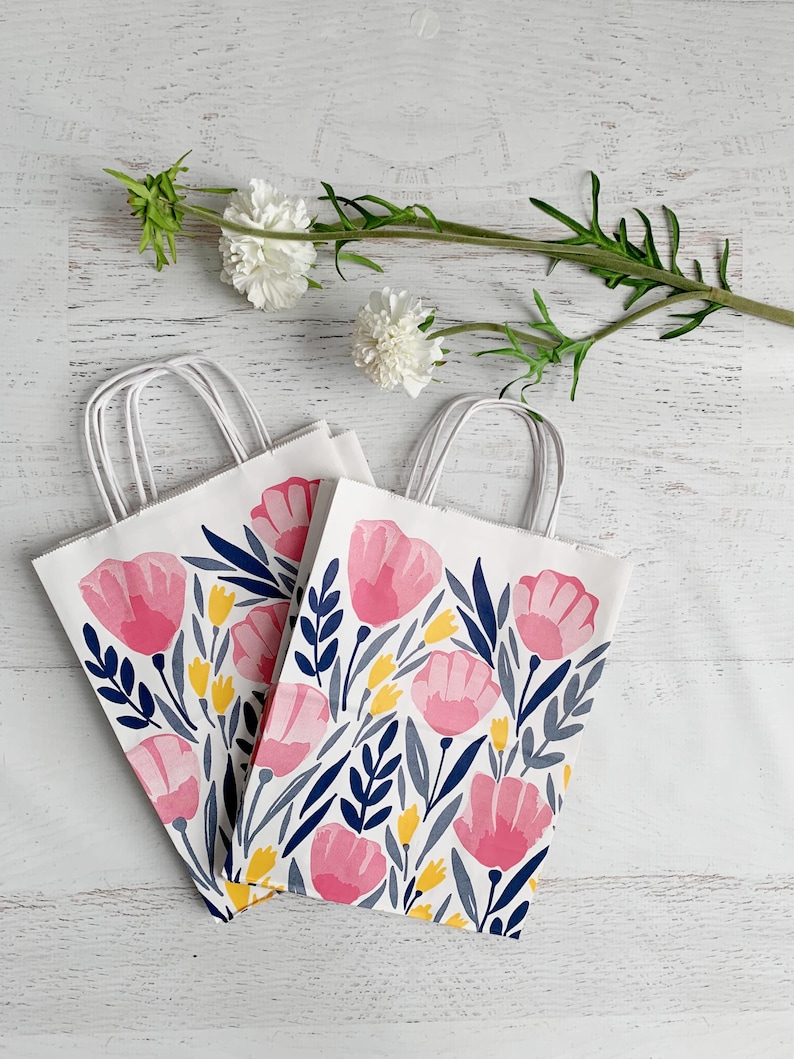 May include: Two white paper gift bags with a floral design featuring pink tulips, navy blue leaves, and yellow accents. The bags have white handles and are arranged on a white wooden surface. A sprig of white flowers is in the background.