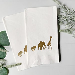 May include: Two white paper napkins with a gold silhouette of a giraffe, elephant, and three smaller animals. The napkins are folded in half and are on a white surface.