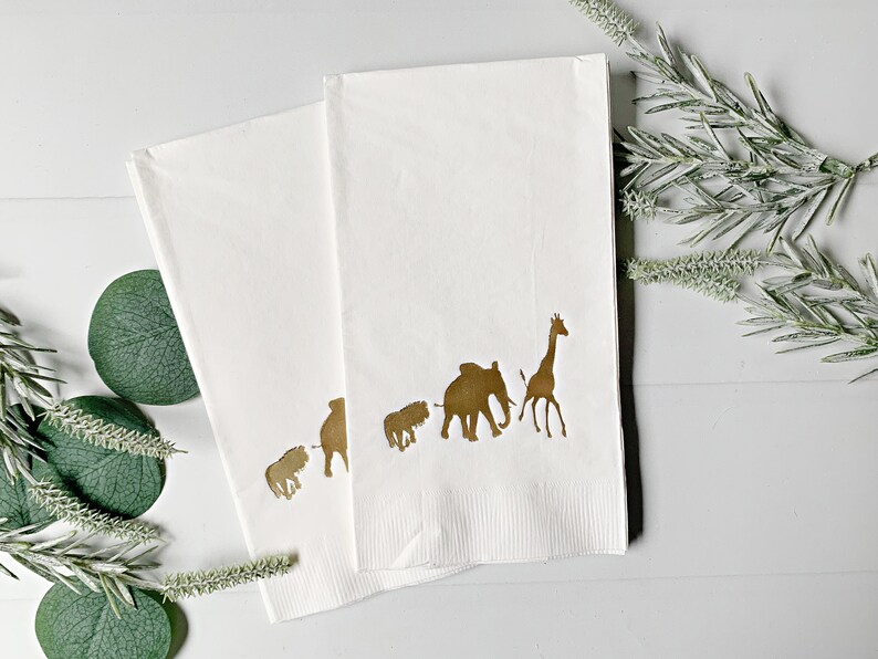 May include: Two white paper napkins with a gold silhouette of a giraffe, elephant, and three smaller animals. The napkins are folded in half and are on a white surface.