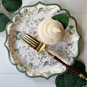 Op de afbeelding: Een decoratieve papieren bord met een geschulpte rand en een bloemmotief in groen en wit. Een cupcake met witte glazuur en een gouden vork met een houten handvat liggen op het bord. Groene bladeren zijn rond het bord gerangschikt.