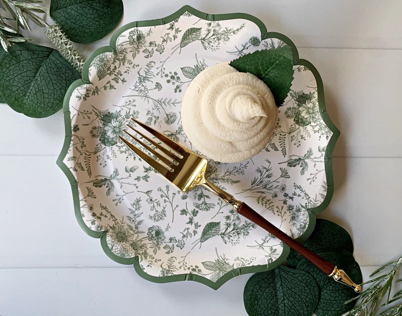 Op de afbeelding: Een decoratieve papieren bord met een geschulpte rand en een bloemmotief in groen en wit. Een cupcake met witte glazuur en een gouden vork met een houten handvat liggen op het bord. Groene bladeren zijn rond het bord gerangschikt.