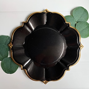 Op de afbeelding: Een zwarte decoratieve schaal met een gouden rand en een geschulpte rand. De schaal is omgeven door groene bladeren op een wit houten oppervlak. De schaal heeft een glanzende afwerking.