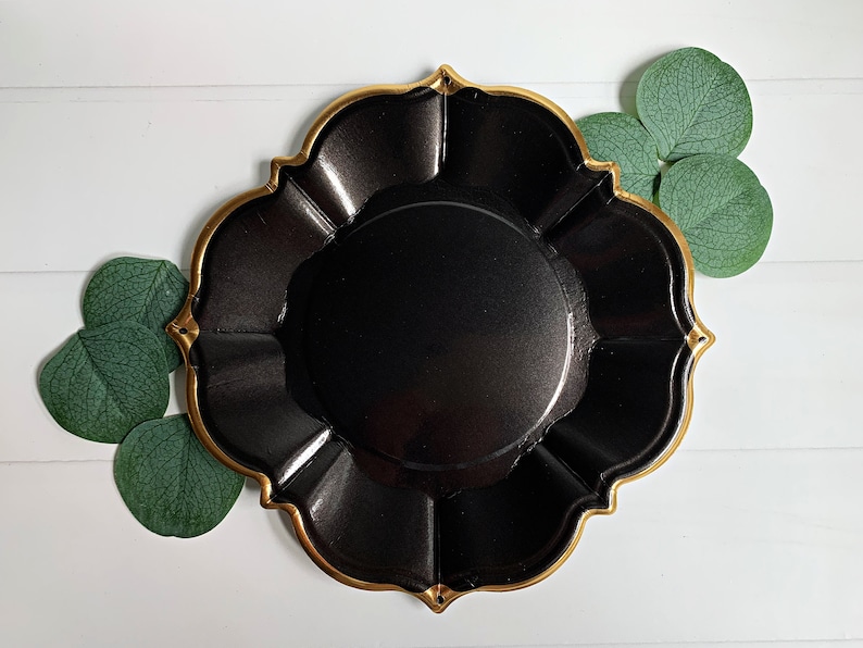 Op de afbeelding: Een zwarte decoratieve schaal met een gouden rand en een geschulpte rand. De schaal is omgeven door groene bladeren op een wit houten oppervlak. De schaal heeft een glanzende afwerking.