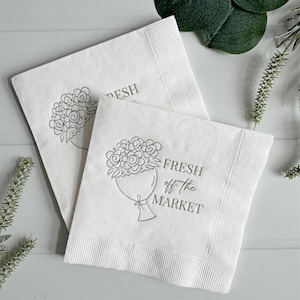 May include: Two white paper napkins with a green floral bouquet design and the text "FRESH off the MARKET". The napkins have a ribbed border and are arranged on a light gray surface.