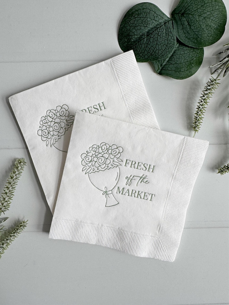 May include: Two white paper napkins with a green floral bouquet design and the text "FRESH off the MARKET". The napkins have a ribbed border and are arranged on a light gray surface.