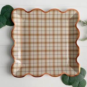 May include: A square paper plate with a plaid pattern in beige, brown, and black. The plate has a scalloped edge with a copper-colored trim. Green leaves and sprigs of greenery are arranged around the plate.