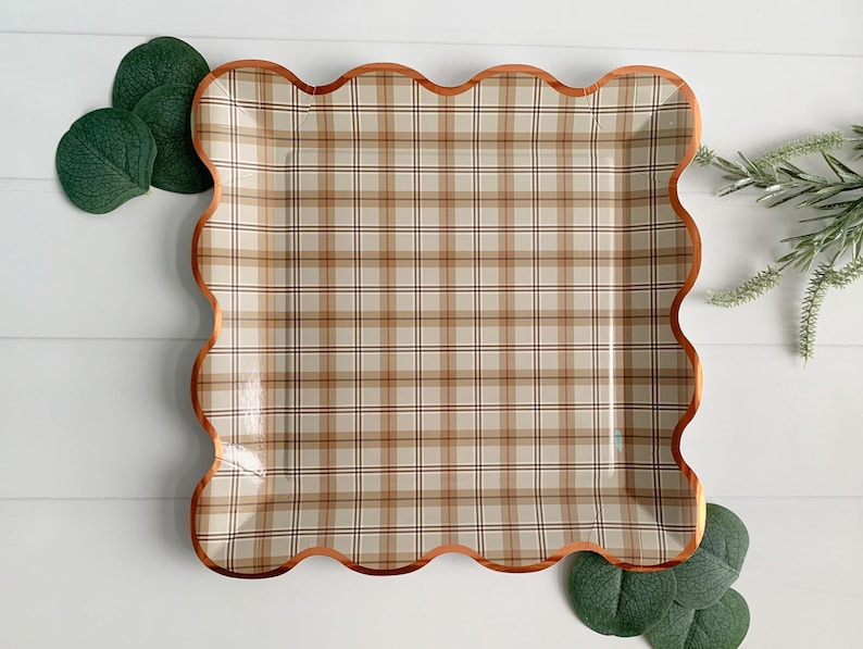 May include: A square paper plate with a plaid pattern in beige, brown, and black. The plate has a scalloped edge with a copper-colored trim. Green leaves and sprigs of greenery are arranged around the plate.