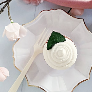 May include: A single cupcake with white frosting and a green leaf on top, sitting on a white scalloped plate with a gold rim. A white plastic fork is also on the plate.