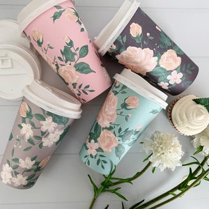 Puede incluir: Cuatro vasos de caf&eacute; de papel con estampado floral y tapas blancas. Los vasos tienen diferentes patrones florales en rosa, gris y azul. Los vasos est&aacute;n dispuestos sobre una superficie de madera blanca con una magdalena y flores blancas.