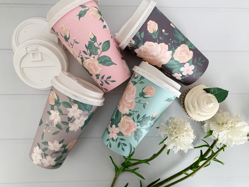 Puede incluir: Cuatro vasos de caf&eacute; de papel con estampado floral y tapas blancas. Los vasos tienen diferentes patrones florales en rosa, gris y azul. Los vasos est&aacute;n dispuestos sobre una superficie de madera blanca con una magdalena y flores blancas.