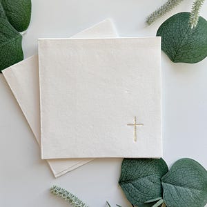 May include: Off-white square cards with a gold cross emblem, stacked on top of each other. The cards are surrounded by green foliage on a white background. The cards are likely for a religious event.