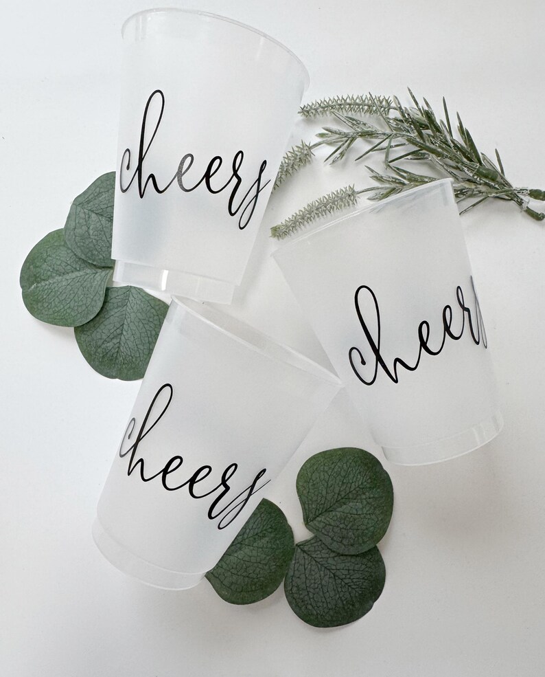 May include: Three frosted plastic cups with the word "cheers" in black script. The cups are arranged with green leaves and sprigs of greenery. The cups are ideal for parties and celebrations.