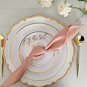 May include: A white dinner plate with a scalloped gold rim and a smaller white salad plate featuring a gold floral line drawing. A dusty pink cloth napkin is knotted on top. Gold-toned flatware includes a fork, knife, and spoon.