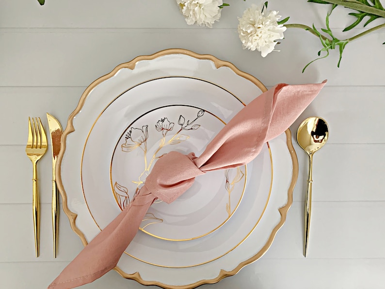 May include: A white dinner plate with a scalloped gold rim and a smaller white salad plate featuring a gold floral line drawing. A dusty pink cloth napkin is knotted on top. Gold-toned flatware includes a fork, knife, and spoon.