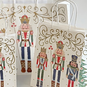 May include: White paper gift bags with illustrations of nutcrackers in various uniforms. The bags have white twisted paper handles and gold decorative flourishes. The nutcrackers are in red, green, blue, and brown. A candy cane is held by one of the nutcrackers.