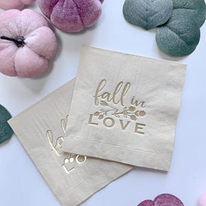May include: Two square beige paper napkins featuring the gold foil text "fall in LOVE" with a small botanical illustration. The napkins are surrounded by several small velvet pumpkins in shades of pink and purple, along with green eucalyptus leaves.