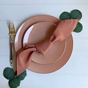 May include: A brown napkin tied in a bow on top of two blush pink plates with a gold rim. The plates are on a white surface with green leaves.