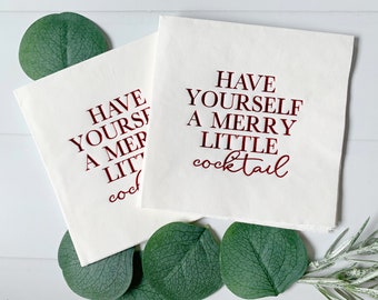May include: Two white napkins with red text that reads "Have yourself a merry little cocktail". The napkins are on a white wooden surface with green leaves.