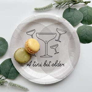 May include: A round, light gray paper plate with a black line drawing of cocktail glasses and the phrase "A tini bit older." Two macarons, one yellow and one green, sit on the plate. Green leaves and sprigs of greenery surround the plate.