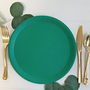 May include: A green paper plate with a gold fork, knife, and spoon set on a white wooden surface. Green eucalyptus leaves are arranged around the plate. The tableware is set for a meal.