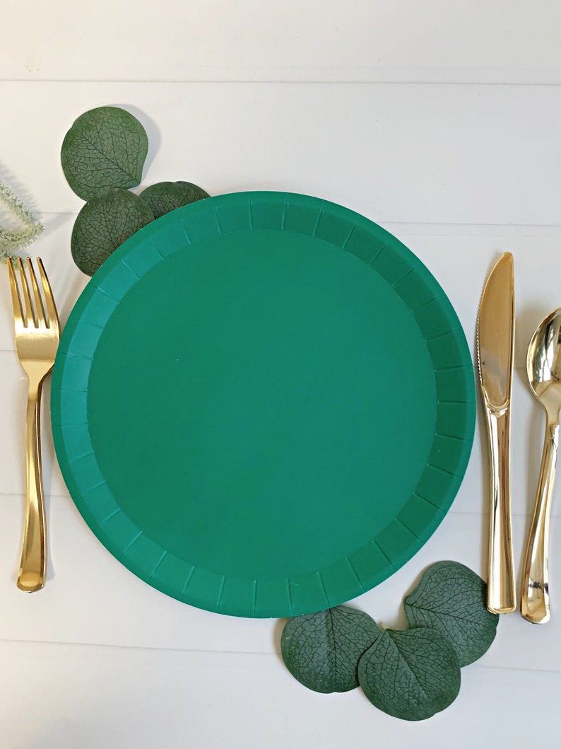 May include: A green paper plate with a gold fork, knife, and spoon set on a white wooden surface. Green eucalyptus leaves are arranged around the plate. The tableware is set for a meal.