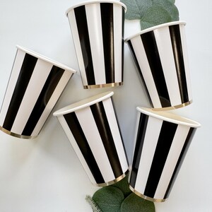 May include: Five disposable paper cups featuring a bold vertical black and white striped pattern. Each cup has a metallic gold rim at the base. The cups are arranged in a circular formation with green eucalyptus leaves tucked between them.