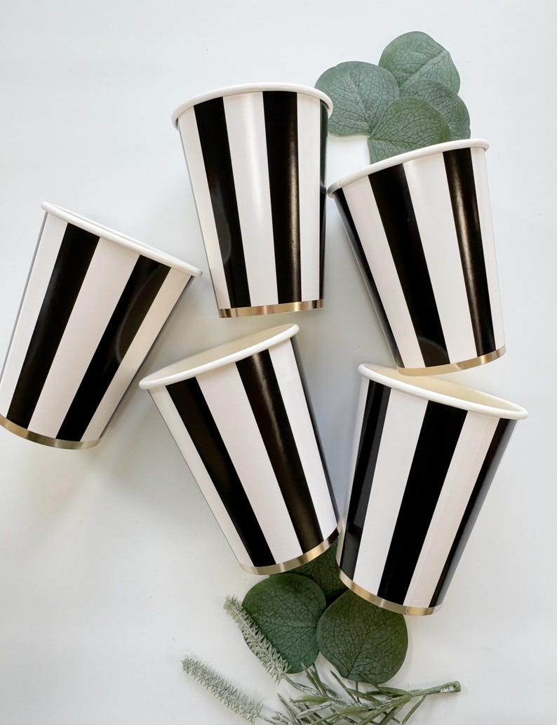 May include: Five disposable paper cups featuring a bold vertical black and white striped pattern. Each cup has a metallic gold rim at the base. The cups are arranged in a circular formation with green eucalyptus leaves tucked between them.