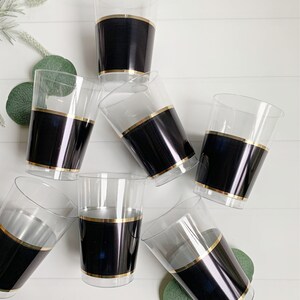 May include: Clear plastic cups with a black band and gold trim. The cups are arranged on a white surface with green foliage. The cups are suitable for serving beverages at parties and events.