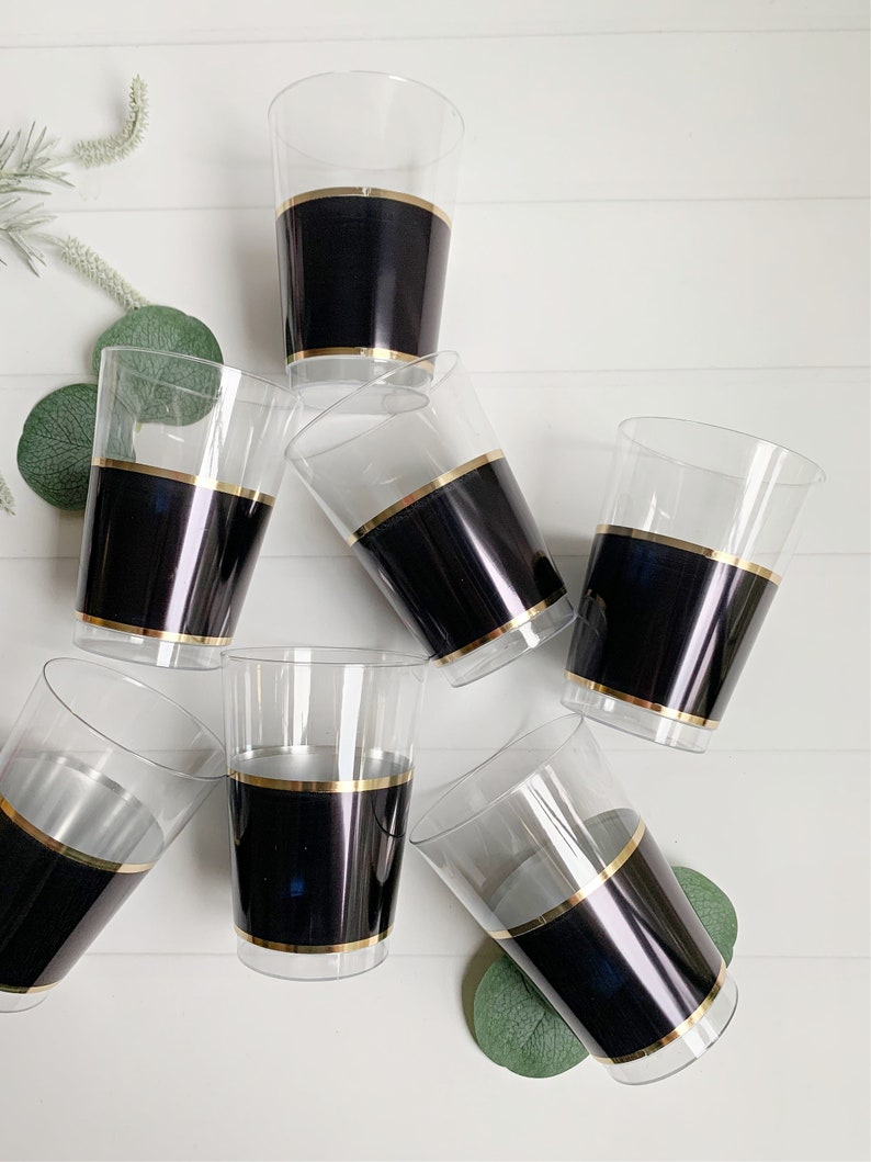 May include: Clear plastic cups with a black band and gold trim. The cups are arranged on a white surface with green foliage. The cups are suitable for serving beverages at parties and events.