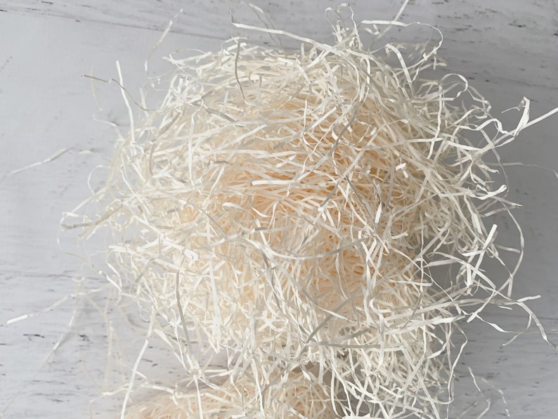 Op de afbeelding: Close-up van een stapel lichtbeige versnipperd papier, gebruikt voor verpakking en demping. Het papier is gerangschikt in een dichte, verwarde massa, met individuele stroken zichtbaar. De achtergrond is een licht, getextureerd oppervlak.