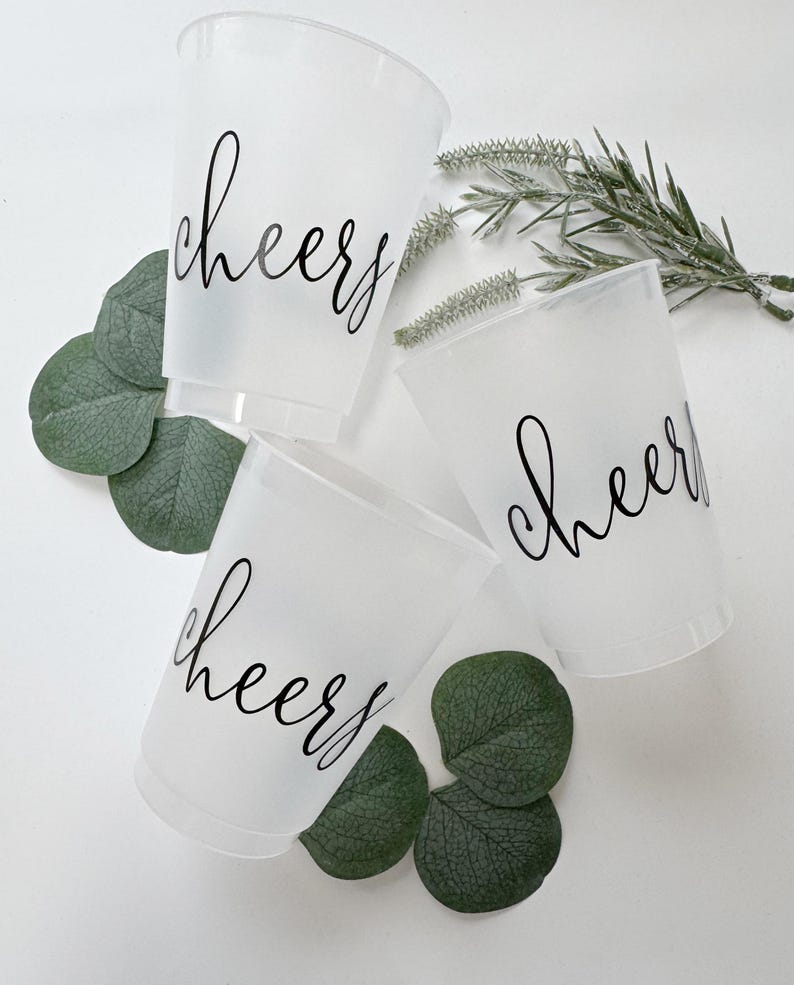 May include: Three translucent frosted plastic cups, each featuring the word cheers printed in black cursive script. The cups are arranged with green eucalyptus leaves and a sprig of greenery tucked between them on a plain white surface.