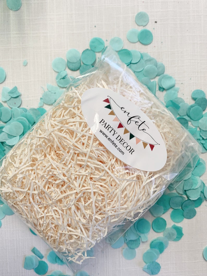 Op de afbeelding: Een doorzichtige plastic zak gevuld met versnipperd papier, met een ovaal wit etiket met de tekst "enfete PARTY DECOR www.enfete.com". Lichtblauwe confetti cirkels zijn verspreid.