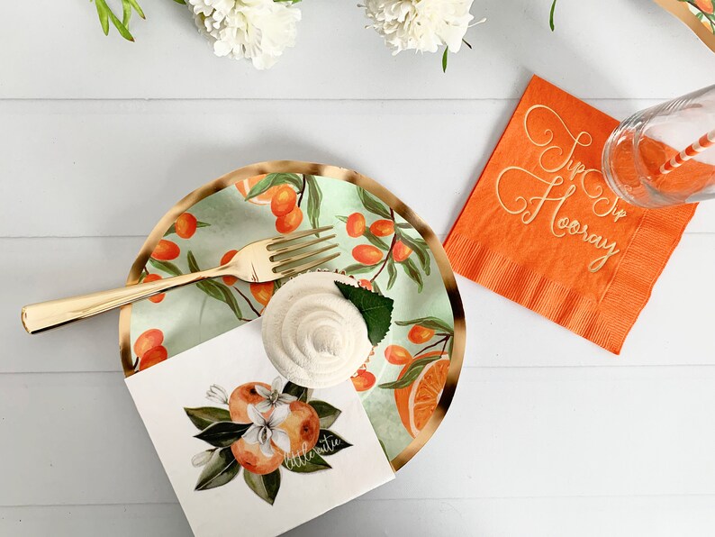 Op de afbeelding: Een feestelijke tafelsetting met een gouden vork, een cupcake en een bord met een sinaasappel- en bladerontwerp. Een oranje servet met de tekst "Hip Hip Hooray" en een glas met een gestreept rietje zijn ook aanwezig. Thema: feest.