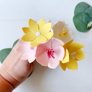 May include: A bouquet of paper flowers in shades of pink, yellow, and white. The flowers are made of paper and have a 3D effect. The flowers are arranged in a hand.