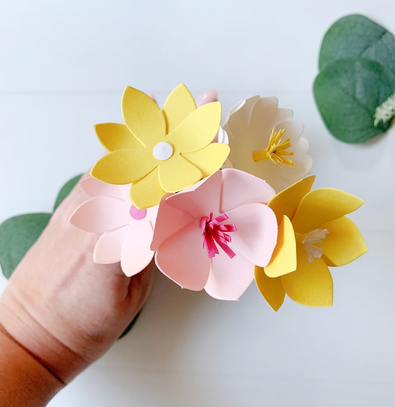 May include: A bouquet of paper flowers in shades of pink, yellow, and white. The flowers are made of paper and have a 3D effect. The flowers are arranged in a hand.