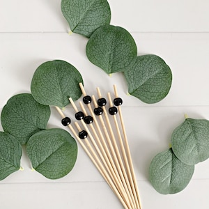 May include: A collection of bamboo skewers topped with black beads, arranged with green eucalyptus leaves. The skewers are natural wood color, and the leaves are a deep green. The arrangement is on a white surface.